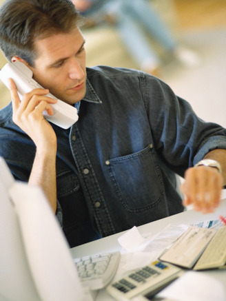 Man Sitting at Desk, on Phone While Working on Computer